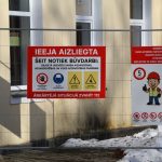 Pie metāla žoga pie dzeltenas ēkas ar logiem, kurā notiek būvdarbi, piestiprinātas divas informatīvas zīmes. Kreisajā sarkanajā zīmē teikts "IEEJA AIZLIEGTA ŠEIT NOTIEK BŪVDARBI: OBJEKTĀ JĀIEVĒRO DARBA AIZSARDZĪBAS, UGUNSDROŠĪBAS UN VIDES AIZSARDZĪBAS PRASĪBAS! ĀRKĀRTĒJĀ SITUĀCIJĀ ZVANĪT 112" ar ikonām par ieejas aizliegumu, obligātu ķiveres lietošanu, vispārēju bīstamību un paceltu kravu. Labajā zīmē, kas ir balta ar sarkanu virsrakstu, rakstīts "UZMANĪBU! KRAVAS AUTOMOBIĻU VADĪTĀJIEM ATRODOTIES ĀRPUS TRANSPORTLĪDZEKĻA JĀLIETO: ATTENTION! WHEN OUTSIDE THE VEHICLE TRUCK DRIVERS MUST WEAR:", ar piktogrammu, kas attēlo celtnieku un uzskaita obligāto aizsargaprīkojumu (ķivere, veste, zābaki), ātruma ierobežojumu 5 km/h, un citas drošības zīmes. Priekšplānā uz zemes ir sniegs.