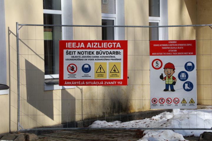 Pie metāla žoga pie dzeltenas ēkas ar logiem, kurā notiek būvdarbi, piestiprinātas divas informatīvas zīmes. Kreisajā sarkanajā zīmē teikts "IEEJA AIZLIEGTA ŠEIT NOTIEK BŪVDARBI: OBJEKTĀ JĀIEVĒRO DARBA AIZSARDZĪBAS, UGUNSDROŠĪBAS UN VIDES AIZSARDZĪBAS PRASĪBAS! ĀRKĀRTĒJĀ SITUĀCIJĀ ZVANĪT 112" ar ikonām par ieejas aizliegumu, obligātu ķiveres lietošanu, vispārēju bīstamību un paceltu kravu. Labajā zīmē, kas ir balta ar sarkanu virsrakstu, rakstīts "UZMANĪBU! KRAVAS AUTOMOBIĻU VADĪTĀJIEM ATRODOTIES ĀRPUS TRANSPORTLĪDZEKĻA JĀLIETO: ATTENTION! WHEN OUTSIDE THE VEHICLE TRUCK DRIVERS MUST WEAR:", ar piktogrammu, kas attēlo celtnieku un uzskaita obligāto aizsargaprīkojumu (ķivere, veste, zābaki), ātruma ierobežojumu 5 km/h, un citas drošības zīmes. Priekšplānā uz zemes ir sniegs.