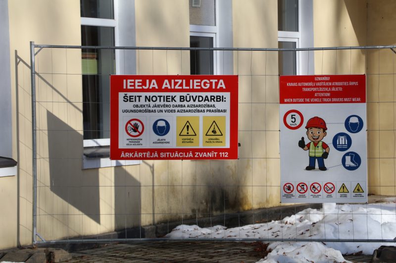 Pie metāla žoga pie dzeltenas ēkas ar logiem, kurā notiek būvdarbi, piestiprinātas divas informatīvas zīmes. Kreisajā sarkanajā zīmē teikts "IEEJA AIZLIEGTA ŠEIT NOTIEK BŪVDARBI: OBJEKTĀ JĀIEVĒRO DARBA AIZSARDZĪBAS, UGUNSDROŠĪBAS UN VIDES AIZSARDZĪBAS PRASĪBAS! ĀRKĀRTĒJĀ SITUĀCIJĀ ZVANĪT 112" ar ikonām par ieejas aizliegumu, obligātu ķiveres lietošanu, vispārēju bīstamību un paceltu kravu. Labajā zīmē, kas ir balta ar sarkanu virsrakstu, rakstīts "UZMANĪBU! KRAVAS AUTOMOBIĻU VADĪTĀJIEM ATRODOTIES ĀRPUS TRANSPORTLĪDZEKĻA JĀLIETO: ATTENTION! WHEN OUTSIDE THE VEHICLE TRUCK DRIVERS MUST WEAR:", ar piktogrammu, kas attēlo celtnieku un uzskaita obligāto aizsargaprīkojumu (ķivere, veste, zābaki), ātruma ierobežojumu 5 km/h, un citas drošības zīmes. Priekšplānā uz zemes ir sniegs.