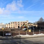 Nojaukšanas laukums ar dzeltenu ekskavatoru, gruvešu kaudzēm un daļēji sniegotu zemi. Fonā redzamas vecas, daļēji nojauktas ēkas un jauna rūpnīcas ēka ar dūmiem. Uz žoga ir zīme ar uzrakstu "RUBATE".