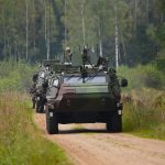 Vairāki maskēšanās krāsas militārie bruņutransportieri brauc kolonnā pa grants ceļu mežainā apvidū. Priekšgalā esošajā transportierī redzams karavīrs.