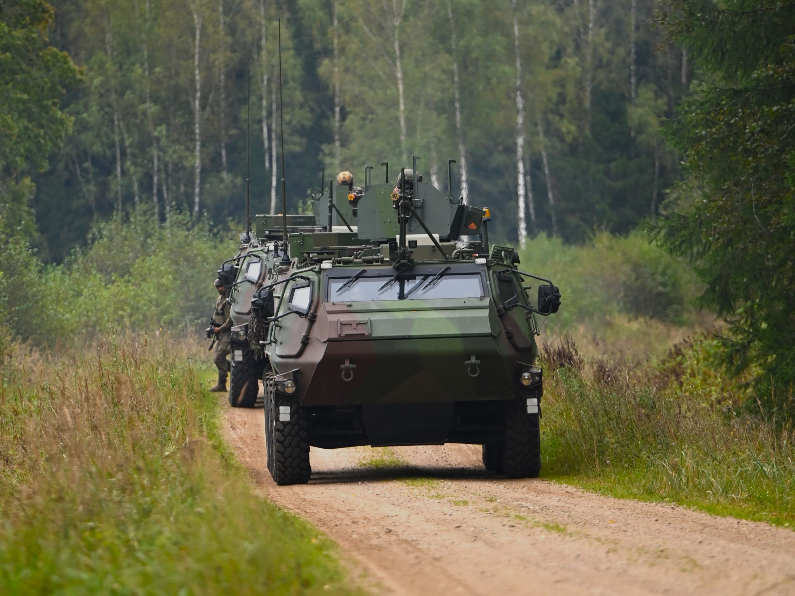 Vairāki maskēšanās krāsas militārie bruņutransportieri brauc kolonnā pa grants ceļu mežainā apvidū. Priekšgalā esošajā transportierī redzams karavīrs.