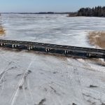 Garš, zems tilts ar tumšu virsmu un gaišiem balstiem stiepjas pāri plašam, aizsalušam ezeram, uz kura ledus redzamas daudzas pēdas. Ezera krastos aug koki un brūnas niedres zem skaidrām zilām debesīm.