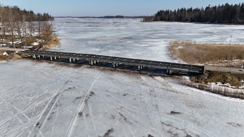 Garš, zems tilts ar tumšu virsmu un gaišiem balstiem stiepjas pāri plašam, aizsalušam ezeram, uz kura ledus redzamas daudzas pēdas. Ezera krastos aug koki un brūnas niedres zem skaidrām zilām debesīm.
