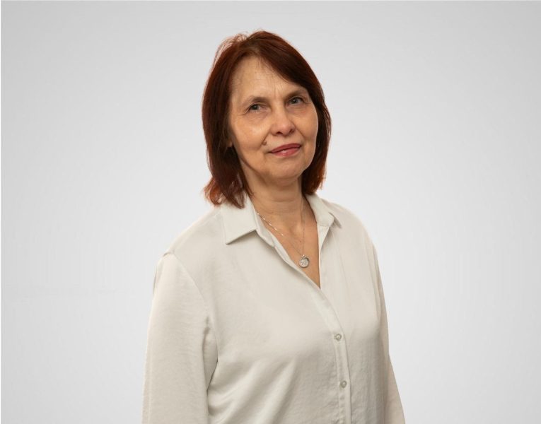 Attēlā sieviete tumšiem matiem un baltā blūzē.