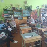 Spilgti zaļā klasē vairāki pieaugušie sēž kreisajā pusē, daži ar papīriem rokās, un vēro bērnus, kuri ir iesaistīti aktivitātēs pie koka galdiem attēla labajā pusē. Fonā redzami plaukti ar mapēm, grāmatām un dekorācijām.