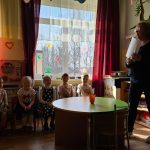 Klasē ar gaiši zaļām sienām pieaugušie sēž krēslos un novēro bērnus, kuri pie koka galdiem aktīvi iesaistījušies mācību aktivitātēs. Aizmugurē redzami plaukti ar mapēm un dekorācijām.