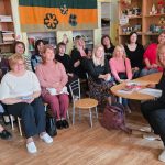 Klases telpa ar zaļām sienām, kurā pieaugušās sievietes sēž kreisajā pusē, bet zēni sēž pie koka galdiem labajā pusē, nodarbojoties ar kādām aktivitātēm. Fonā redzami koka plaukti ar mapēm, grāmatām un dekoratīviem augiem, kā arī sienas dēlis, kas rotāts ar krāsainiem papīra ziediem.