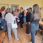 Klases telpa ar gaiši zaļām sienām, kurā pieaugušie sēž kreisajā pusē, vērojot bērnus, kas aktīvi darbojas pie koka galdiem. Bērni ir iesaistīti praktiskās nodarbībās ar papīriem un maziem priekšmetiem. Aizmugurē redzami plaukti ar grāmatām un dekorācijām.