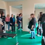 Klase ar gaiši zaļām sienām, kurā pieaugušie sēž kreisajā pusē, bet bērni darbojas pie koka galdiem labajā pusē. Telpas fonā ir plaukti ar grāmatām un mapēm.