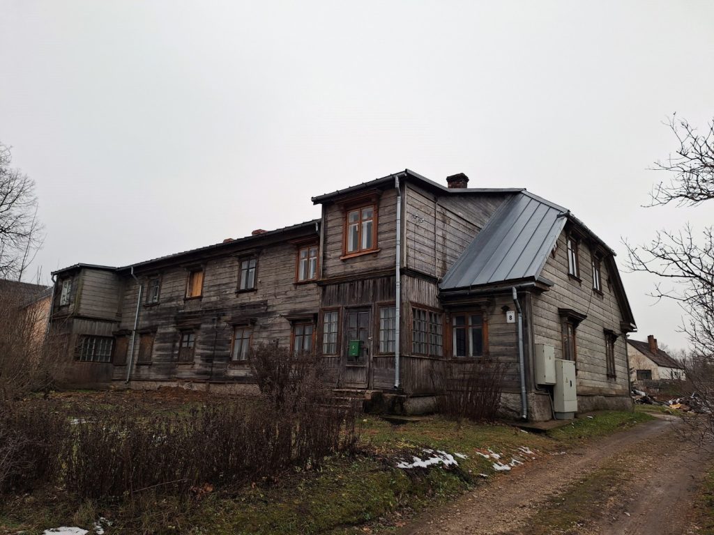 Veclaicīga, divstāvu koka māja ar tumšiem, nodilušiem dēļiem un pelēku metāla jumtu zem apmākušām debesīm. Uz mājas pie ieejas redzama plāksnīte ar numuru "8". Priekšplānā ir zemes ceļš un aizaugusi veģetācija.