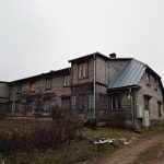 Veclaicīga, divstāvu koka māja ar tumšiem, nodilušiem dēļiem un pelēku metāla jumtu zem apmākušām debesīm. Uz mājas pie ieejas redzama plāksnīte ar numuru "8". Priekšplānā ir zemes ceļš un aizaugusi veģetācija.