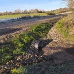 Grants ceļš ar caurteku, no kuras ūdens plūst grāvī. Apsarmojis lauks un koki fonā saulainā dienā.