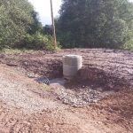 Betona aka ar melnu gofrētu drenāžas cauruli, kas ieplūst ūdenī uz dubļainas zemes ar granti. Fonā redzami koki un koka stabs.