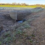 Betona aka ar gofrētu cauruli atrodas izraktā zemē ar ūdens peļķi. Apkārtne ir apsarmojusi, fonā zaļš lauks un koki.