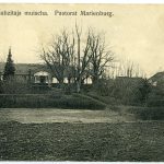 ALUKSNES MAHZITAJA MUISCHA. Pastorat Marienburg. Vēsturiska sēpijas toņa fotogrāfija attēlo lauku ainavu ar vairākām ēkām, tostarp garu māju ar kolonnu portiku, ko ieskauj kaili koki un aramzeme priekšplānā.