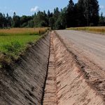 Garš, tukšs meliorācijas grāvis atdala grants ceļu no zaļa lauka. Tālumā redzami koki un zilas debesis.