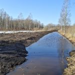 Gara, šaura ūdens grāvis agrā pavasarī, ar izraktas tumšas zemes valni kreisajā pusē. Tālumā pie grāvja redzams dzeltens ekskavators. Abos krastos kaili koki un brūna zāle, vietām vēl sniegs, zem zilām debesīm.