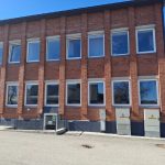 Divstāvu ķieģeļu ēka ar daudziem baltiem logiem saulainā dienā. Pie ēkas pamatiem ir ieeja pagrabstāvā, virs kuras redzama zaļa uzlīme "Patvertne".