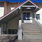 Gaišas ēkas ieeja ar plašām kāpnēm, kas ved uz pelēkām durvīm zem sarkanbrūnas jumta nojumes. Kreisajā pusē ir bruģēta piebrauktuve ar nokāpienu uz pagrabstāva ieeju, kur pie sienas ir zaļa zīme ar uzrakstu "Patvertne".