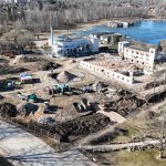 Skats no putna lidojuma uz plašu nojaukšanas un būvniecības sagatavošanas laukumu pie daļēji aizsaluša ezera. Laukumā redzamas nojauktu ēku drupas, būvgružu kaudzes un vairāki ekskavatori ar pašizgāzējiem. Dažas neskartas ēkas atrodas laukuma malās. Apkārtnē ir koki bez lapām un pilsētas apbūve.