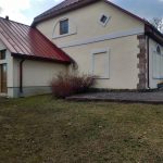 Gaiši dzeltena ēka ar sarkanu metāla jumtu un brūnām durvīm kreisajā pusē. Sienā ir arkveida logs, rombveida logs un tukšs taisnstūrveida rāmis. Stūris dekorēts ar sarkanbrūniem akmeņiem. Priekšplānā zāle un bruģēta taka gar ēkas pamatiem.