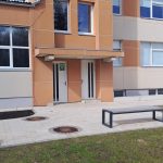 Ēkas ieeja ar divām baltām durvīm. Uz kreisajām durvīm ir zaļa zīme ar uzrakstu "Patvertne". Priekšplānā bruģēts laukums ar soliņu, atkritumu urnu un diviem velosipēdiem.