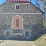 Veca, masīva akmens ēka ar sarkanu dakstiņu jumtu, divām arku durvīm zemes līmenī un aizdarinātu arku logu virs tām. Priekšplānā bruģēts piebraucamais ceļš, fonā – mazāka māja un kaili koki zem zilas debess.