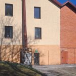 Divstāvu ēka ar gaiši bēšu apmestu daļu kreisajā pusē un sarkanu ķieģeļu daļu labajā pusē. Uz apmestās sienas, kurā ir četri logi, krīt koku ēnas. Pa apmetušās sienas vidu stiepjas brūna caurule, kas ved uz lielu baltu maisu un mazām koka durvīm. Ķieģeļu sienā ir brūnas durvis. Fonā redzami kaili koki, zāļains pakalns un citas ēkas zem skaidri zilām debesīm.