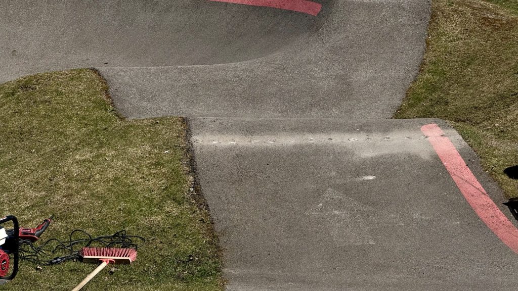 Skats no augšas uz pelēku asfalta pumptrasi ar sarkanām līnijām. Uz zāles blakus trasēm redzams sarkans augstspiediena mazgātājs un slota, kas liecina par trases tīrīšanu.