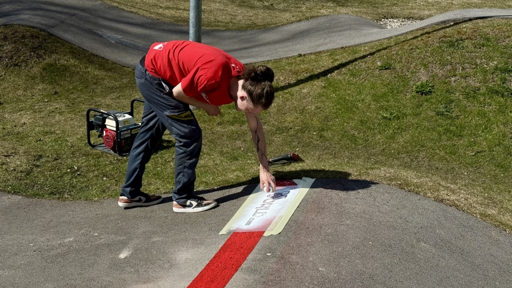 Cilvēks sarkanā kreklā liecas un ar trafareta palīdzību uz asfalta seguma pump track trasē uzkrāso sarkanu logo "Scoop.lv" un "scoop.lv". Blakus uz zāles atrodas portatīvais ģenerators.