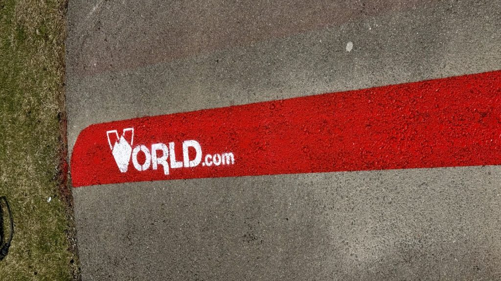 Asfalta segums ar sarkanu līniju, uz kuras baltā krāsā uzkrāsots logotips un teksts "WORLD.com". Kreisajā pusē ir zaļa zāle.