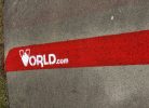 Asfalta segums ar sarkanu līniju, uz kuras baltā krāsā uzkrāsots logotips un teksts "WORLD.com". Kreisajā pusē ir zaļa zāle.