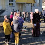 Saulainā dienā pie gaišas ēkas āra laukumā sapulcējusies bērnu grupa un sieviete latviešu tautastērpā. Sieviete ar baltu adītu cepuri, melnu samta jaku un svītrainiem svārkiem stāv priekšplānā pa labi, skatoties pa labi, kamēr bērni ziemas drēbēs stāv apkārt un uzmanīgi vēro.