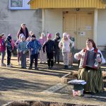 Sieviete tautastērpā spēlē akordeonu, sēžot ārā saulainā dienā. Viņai apkārt stāv bērni un pieaugušie, vērojot priekšnesumu pie ēkas dzeltenajām durvīm. Blakus ir galds ar papīriem un trauku ar krāsainām olām.