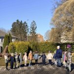 Gados vecāka sieviete tautas tērpā ar sarkanu akordeonu sēž ārā saulainā dienā. Aiz viņas stāv bērni ziemas apģērbā un pieaugušie, kas vēro priekšnesumu pie ēkas uz bruģa.