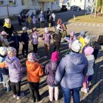 Gados vecāka sieviete tautastērpā sēž un spēlē akordeonu, kamēr viņai pretī stāv bērnu grupa ar pieaugušajiem. Notikums norisinās ārā, saulainā dienā, pie ēkas ar dzeltenām durvīm. Pie sievietes atrodas kaste ar krāsainām bumbiņām.