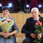 Sieviete un vīrietis sēž blakus uz skatuves, abi turot ziedu pušķus. Sieviete kreisajā pusē smaida, turot krāsainu pušķi, savukārt vīrietis labajā pusē, uzvalkā, turot rozes, izskatās nopietns. Fonā redzamas skatuves gaismas.