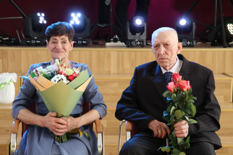 Sieviete un vīrietis sēž blakus uz skatuves, abi turot ziedu pušķus. Sieviete kreisajā pusē smaida, turot krāsainu pušķi, savukārt vīrietis labajā pusē, uzvalkā, turot rozes, izskatās nopietns. Fonā redzamas skatuves gaismas.