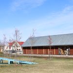 Ziedošs koks ar rozā ziediem priekšplānā, aiz tā – brūna koka ēka ar pelēku metāla jumtu un logu zelminī, zem zilām debesīm ar baltiem mākoņiem.