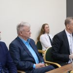 Trīs vīrieši uzvalkos un sieviete baltā žaketē sēž pie galda, uzmanīgi klausoties. Uz galda redzama krūka ar citronūdeni un uzkodas.