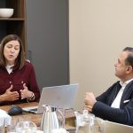 Sieviete tumši sarkanā kreklā runā un žestikulē pie konferenču galda, blakus sēdošais vīrietis tumšā žaketē uzmanīgi klausās. Uz galda redzami portatīvie datori un dzērieni.