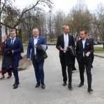 Cilvēku grupa, tostarp vīrieši uzvalkos un sievietes, iet pa āra ceļu un sarunājas. Daži vīrieši rokās tur jakas. Fonā redzamas ēkas un koki bez lapām.