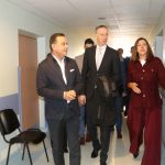 Trīs cilvēki, divi vīrieši un sieviete, staigā pa gaišu medicīnas iestādes koridoru. Sieviete labajā pusē, ģērbusies bordo krāsā, žestikulē ar roku, kamēr vīrieši viņai blakus klausās.