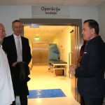 Trīs vīrieši, tostarp viens baltā halātā, sarunājas slimnīcas koridorā. Virs durvīm, kas ved uz spoži apgaismotu gaiteni ar medicīniskiem ratiņiem, ir uzraksts "Operāciju nodaļa".