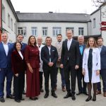 Vienpadsmit cilvēku (septiņi vīrieši, četras sievietes) grupa, tostarp daži medicīnas darbinieki baltos halātos un citi uzvalkos, stāv ārpusē pie ēkas, iespējams, slimnīcas. Labajā pusē redzama virziena zīme ar norādēm: "Administrācija", "Laboratorija", "Bērnu nodaļa", "Ambulatorā nodaļa".