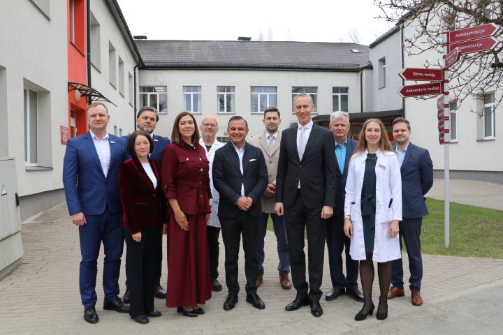 Vienpadsmit cilvēku (septiņi vīrieši, četras sievietes) grupa, tostarp daži medicīnas darbinieki baltos halātos un citi uzvalkos, stāv ārpusē pie ēkas, iespējams, slimnīcas. Labajā pusē redzama virziena zīme ar norādēm: "Administrācija", "Laboratorija", "Bērnu nodaļa", "Ambulatorā nodaļa".