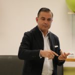 Vīrietis tumšā žaketē un baltā kreklā žestikulē ar rokām, rādot ar labās rokas rādītājpirkstu un turot atvērtu kreiso plaukstu. Fonā redzama gaiši zaļa vingrošanas bumba un tumšs paklājiņš ar daļēji redzamu uzrakstu "RX".