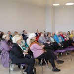 Cilvēku grupa, tostarp trīs vīrieši uzvalkos un sieviete baltā virsvalkā, sēž pie galda sanāksmes telpā, klausoties kādam runātājam. Uz galda ir ūdens krūze ar citroniem un uzkodas.