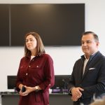 Četri cilvēki, trīs vīrieši un viena sieviete, sēž pie galda sanāksmes telpā, klausoties vai vērojot. Vīrieši tērpti uzvalkos, sieviete baltā virsvalkā. Uz galda ir ūdens krūze ar citronu un uzkodas.